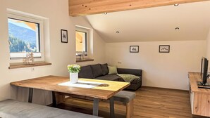 Una habitación moderna y bien iluminada, con una mesa de madera, un banco y un sofá. Dos ventanas ofrecen luz natural y vistas a un área boscosa.