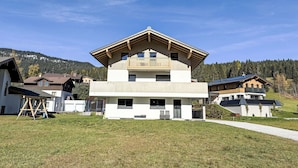 Una casa moderna de dos pisos con balcón, rodeada por un jardín cubierto de césped y un cerro boscoso.