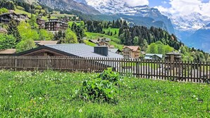 Un paisaje montañoso con una cerca de madera, campos verdes y edificios con techos inclinados.