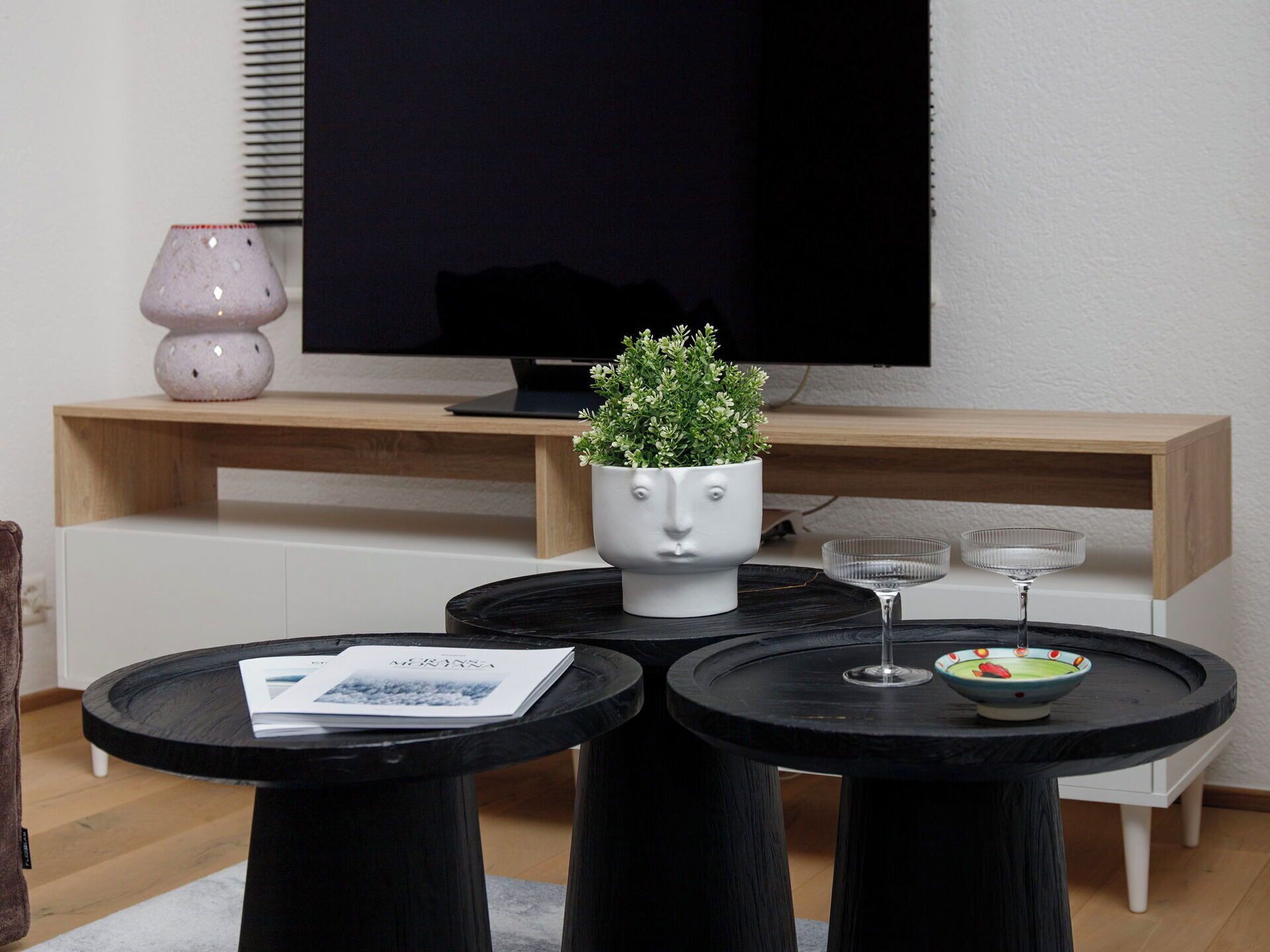 Ett modernt vardagsrum med en tv, en unik blomkruka formad som ett ansikte och två runda bord med drycker på.