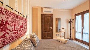 Ett hotellrum med en säng, en bänk, en spegel, en klädställning och ett stort fönster med gardiner.