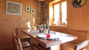 Ein Holzinnenraum mit einer Steinwand, Holzbalkendecke und einem Esstisch mit Stühlen. An der Wand hängen gerahmte Bilder, es gibt ein Fenster mit Vorhängen und eine Hahndekoration.