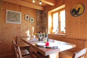 Ein Holzinnenraum mit einer Steinwand, Holzbalkendecke und einem Esstisch mit Stühlen. An der Wand hängen gerahmte Bilder, es gibt ein Fenster mit Vorhängen und eine Hahndekoration.