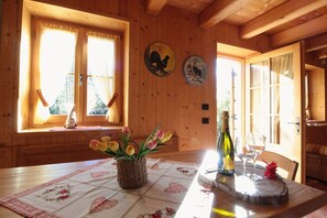 A wooden cabin interior with a dining table, a vase of tulips, a bottle of wine, and two wall-mounted plates depicting chickens.