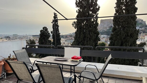 Un balcón con mesa y sillas, con vistas a un paisaje urbano con árboles altos y edificios.