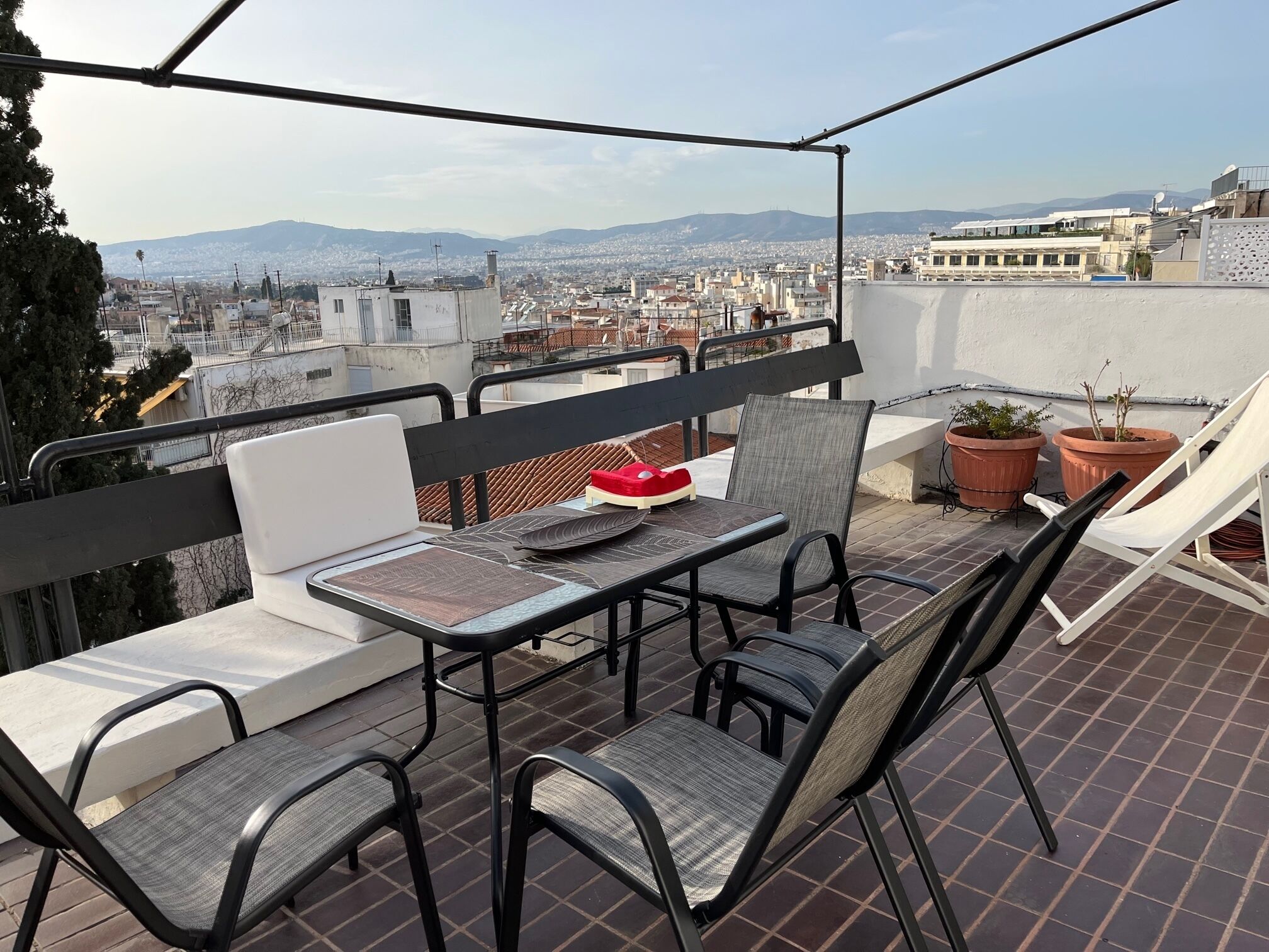An outdoor terrace with a table and chairs, potted plants, and a view of a cityscape.