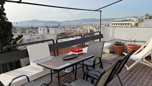 Una terraza al aire libre con una mesa, sillas, plantas en macetas y vistas a un paisaje urbano.