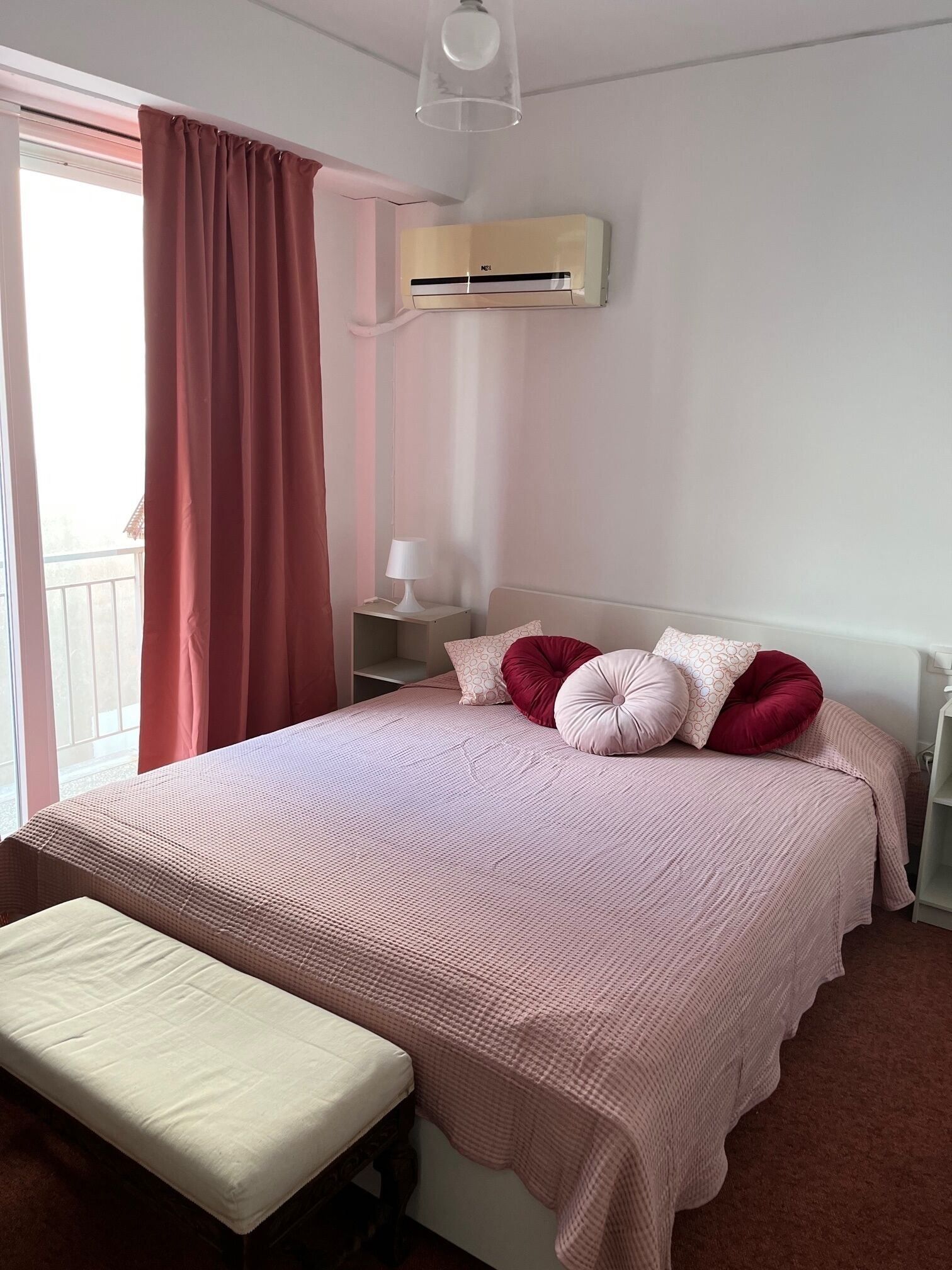 A bedroom with a bed, a bench, a bedside table with a lamp, and an air conditioning unit.