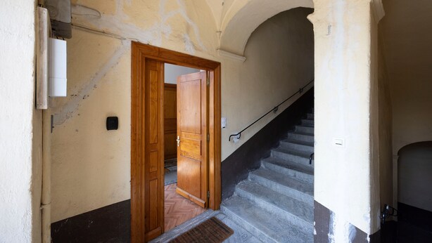 Un pasillo con una puerta de madera que da a una habitación, una escalera con barandilla y un aplique de pared.