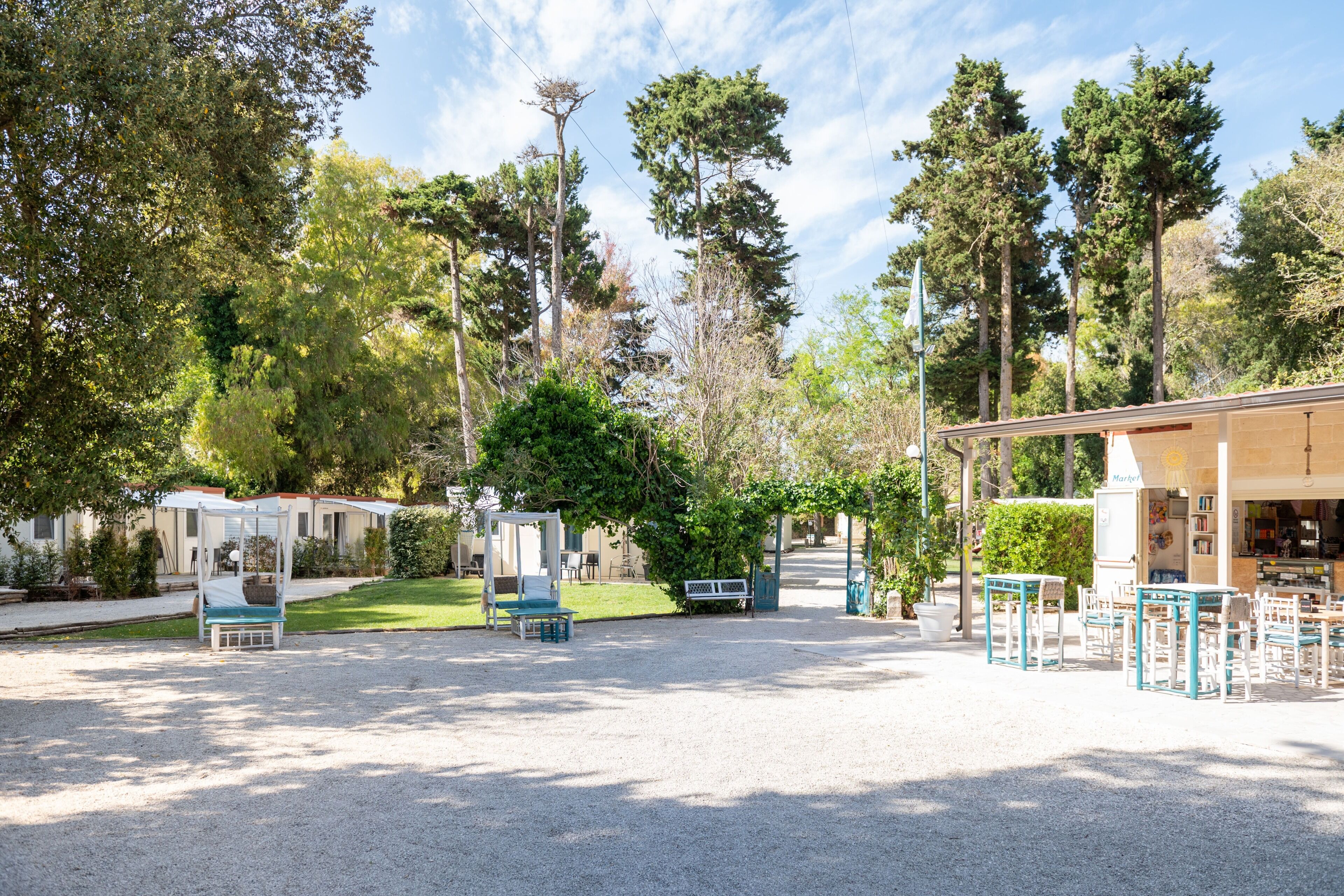 A covered outdoor seating area with tables and chairs, surrounded by trees and a building with a sign.