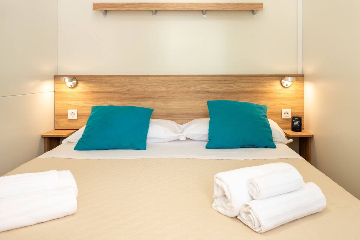A neatly made bed with teal pillows, white bedding, and two rolled towels on the side.