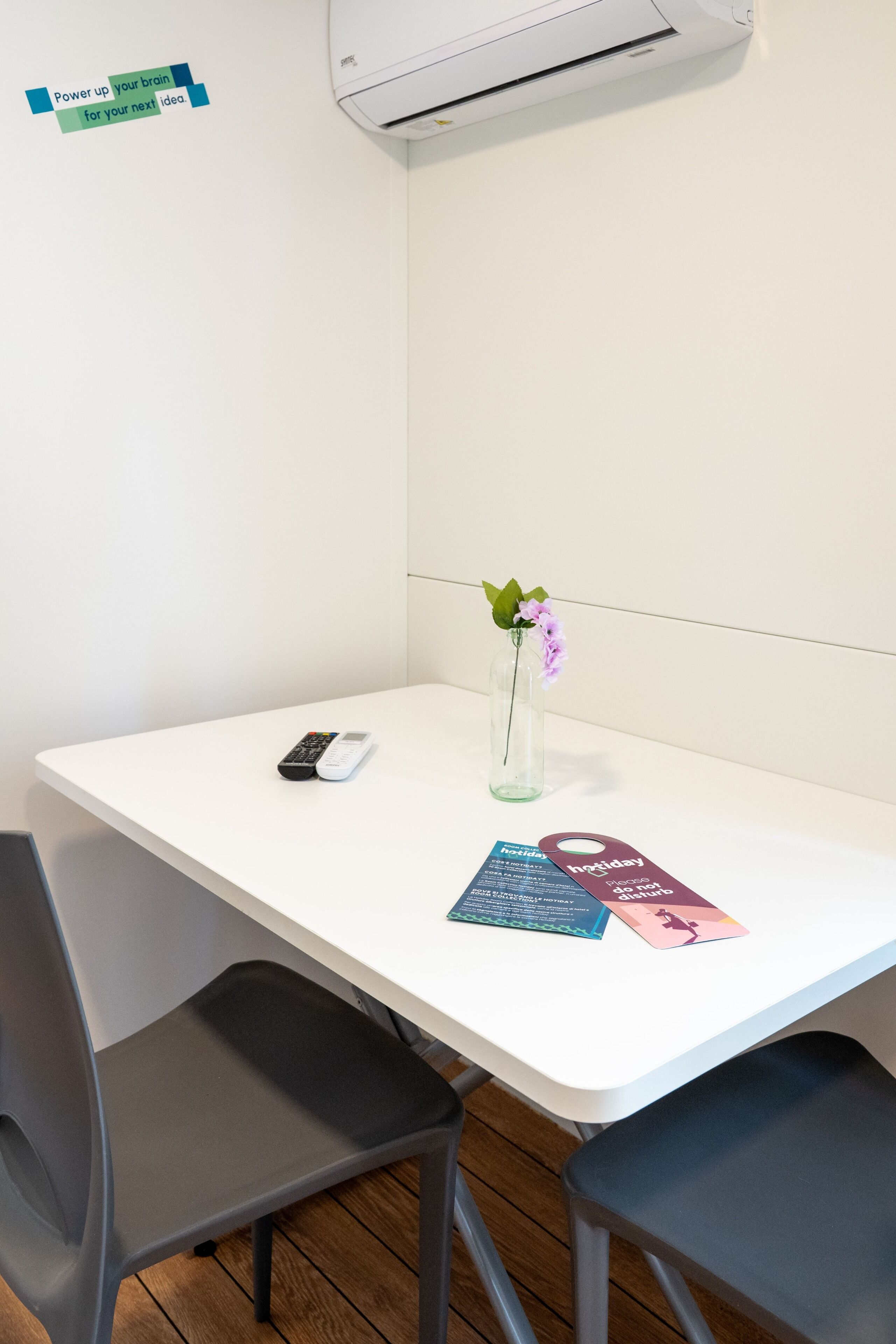 A white table with a remote control, a vase with a flower, and two pamphlets.