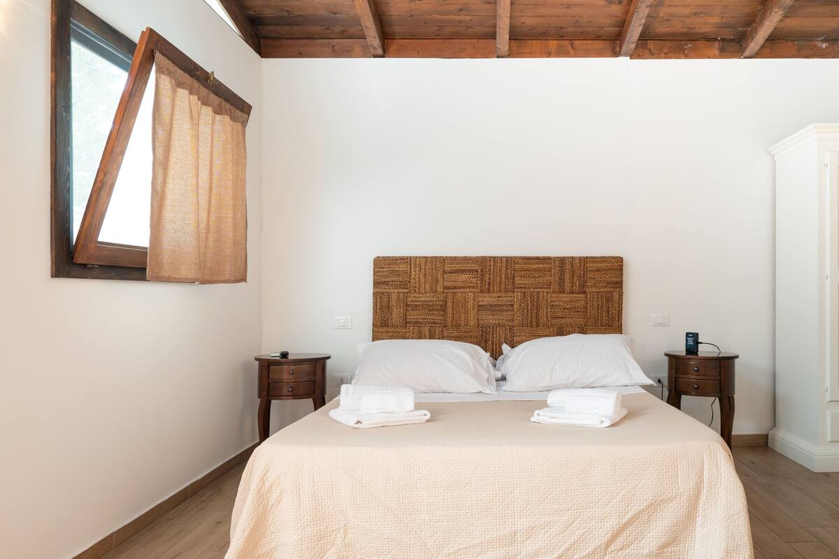 A bedroom with a wooden headboard, two bedside tables, a window with curtains, and a wardrobe.