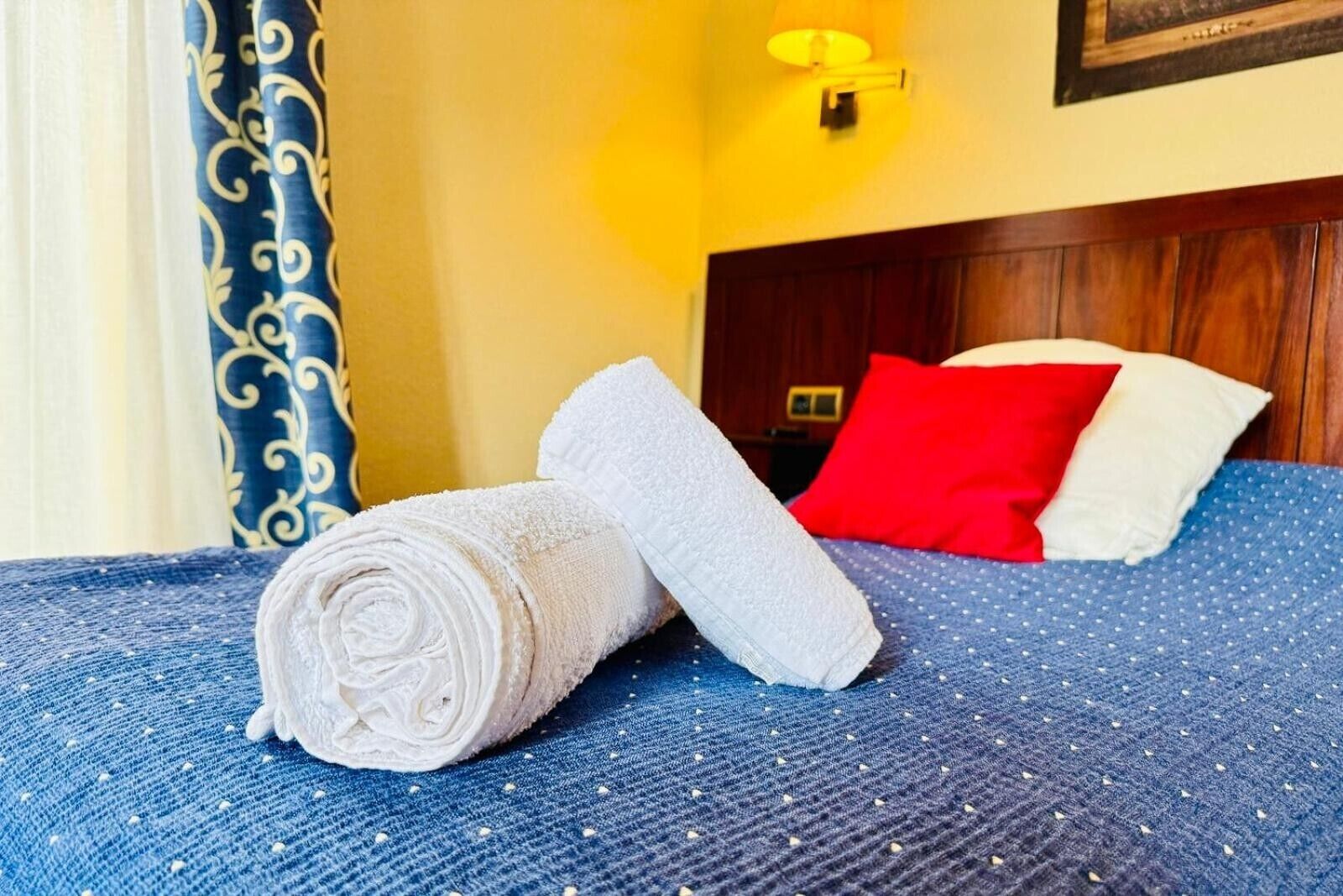 Un lit bien fait, avec une serviette blanche roulée et un oreiller blanc plié, un oreiller rouge et une tête de lit en bois.