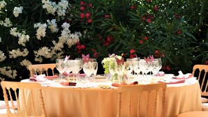 Ein runder Tisch, gedeckt für eine Mahlzeit, mit Stühlen umgeben, inmitten eines üppigen Gartens mit weißen und roten Blumen.
