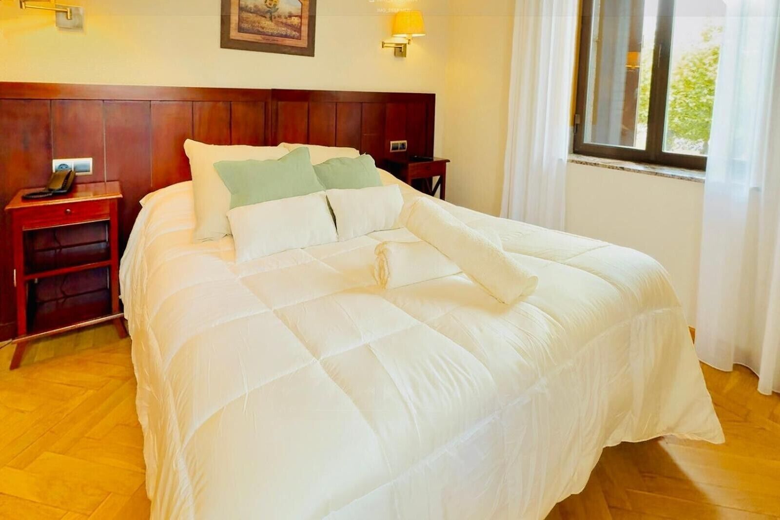 Una cama bien hecha con edredón blanco acolchado, cabecera de madera, mesitas de noche con lámparas y una ventana con cortinas.