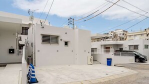 A residential area with white buildings, a black car parked on the street, and a blue trash bin. - New Luxury Home in Koza | Work-Friendly | Free Parking (沖縄県)