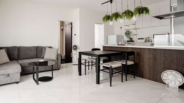 A modern living room with a gray sofa, a dining table with chairs, a ceiling fan, and a kitchen area with a white countertop and hanging plants. - New Luxury Home in Koza | Work-Friendly | Free Parking (沖縄県)