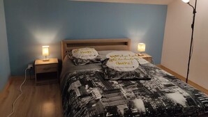 A bedroom with a cityscape bedspread, two bedside lamps, and pillows with text. - Chez Sam - Bed & Breakfast (Villebois)