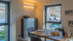 A dining area with a table set for two, a bottle of wine, and a view of a mountain cabin through a window. - MAISON GLENDA - Breathe in the alpine stillness (Courmayeur)