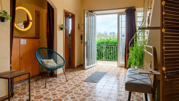 A room with a patterned floor, a chair, a bench, a mirror, and a door leading to a balcony with a view of greenery. - Holiday Home "Villa for 8 Guests – Porte du Ried" with Mountain View, Private Terrace & Wi-Fi (porte du ried)