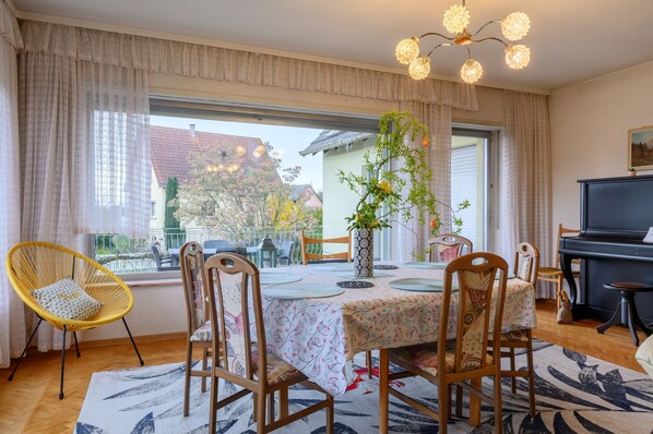 Ein Essbereich mit einem Tisch für vier Personen, Stühlen, einem Klavier und einem großen Fenster mit Vorhängen.