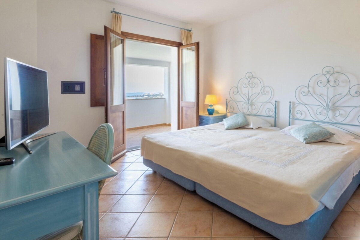 A hotel room with a bed, a desk with a television, a window with a view, and a door leading to an outdoor area.