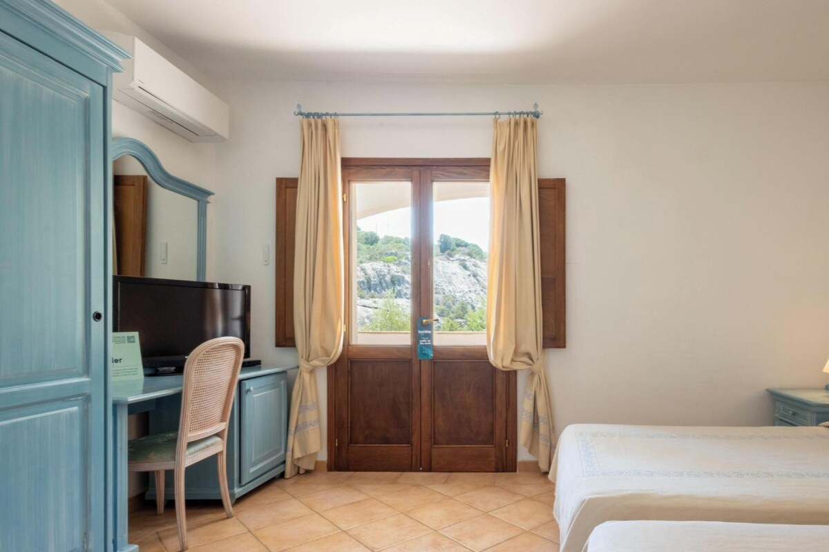 A bedroom with a bed, a desk, a television, and a window with curtains.