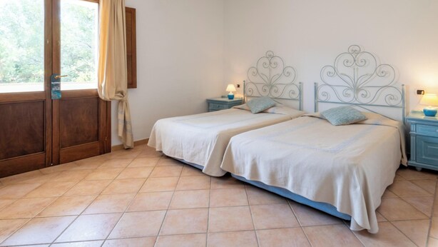 Una cama doble con cabecera metálica, una mesita de noche con lámpara y vista al exterior por una puerta corrediza.