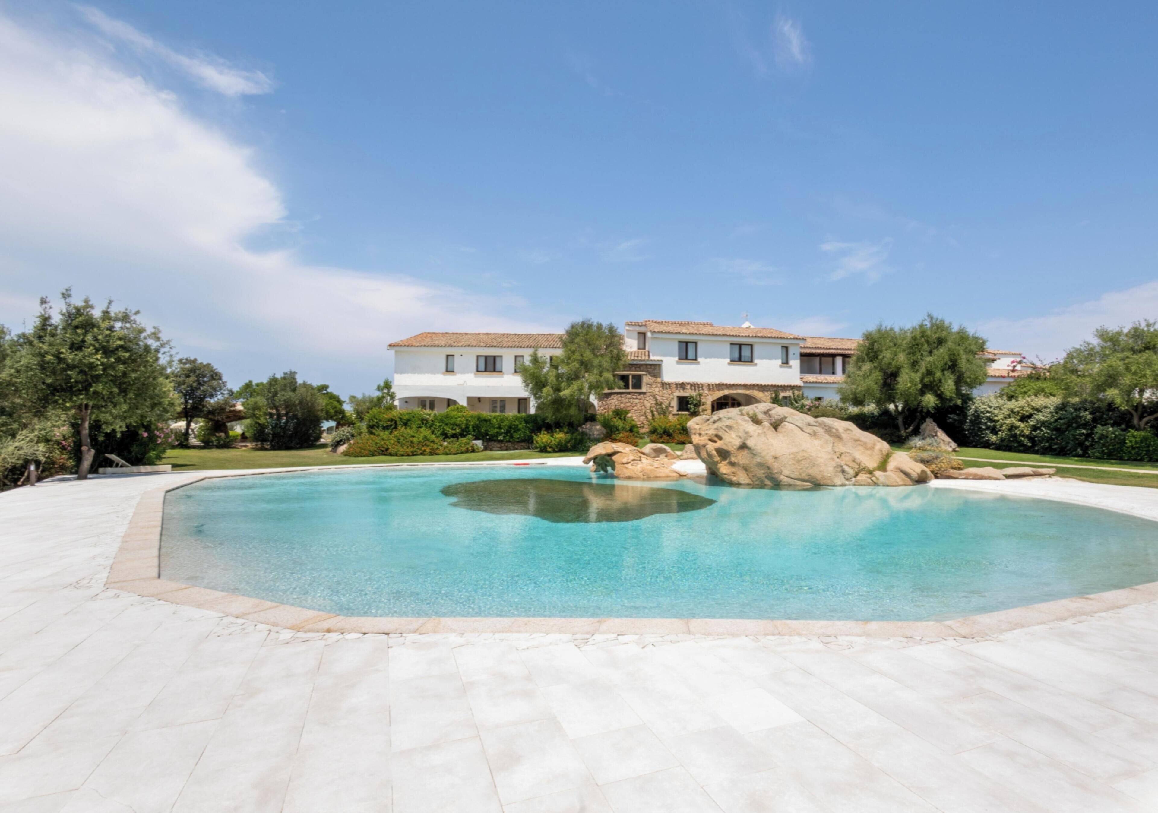 A large, clear swimming pool with a stone border, surrounded by a paved area and a large building with a tiled roof.
