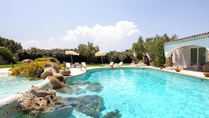 A clear swimming pool with rocks, lounge chairs, and umbrellas. - Hotiday Baja Sardinia Saline (Baja Sardinia)