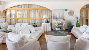 Un salón espacioso con techo alto, mobiliario blanco y amplias ventanas de madera.