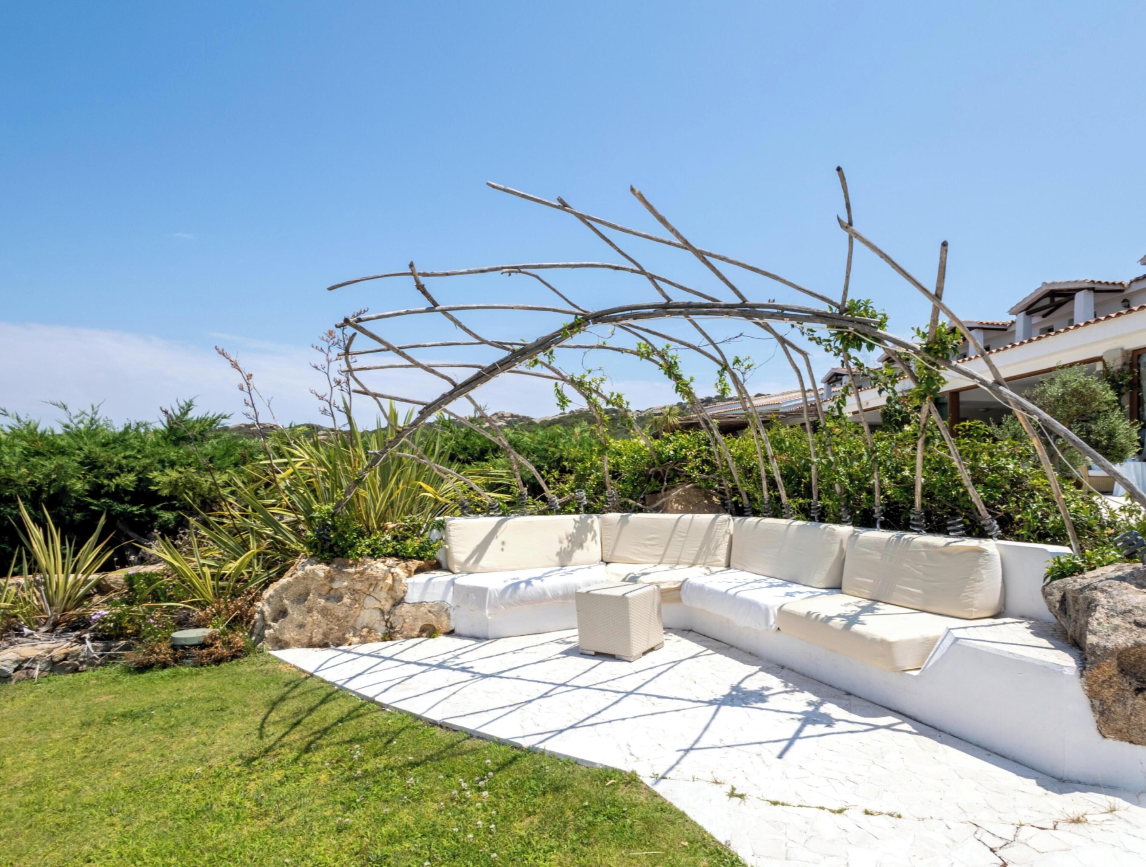 A garden with a white outdoor seating area, surrounded by greenery and a stone wall.
