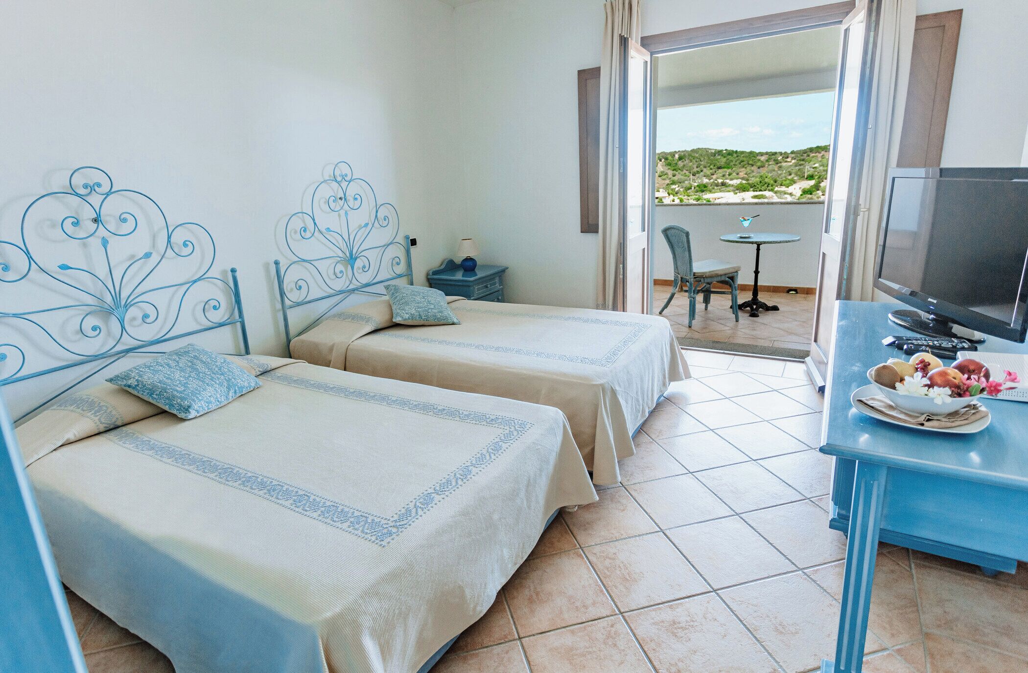 A hotel room with two beds, a TV, a small table with a bowl of fruit, and a balcony with a view.