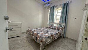 A bedroom with a bed, a dresser, a window with curtains, and an air conditioning unit. - Prime Apartment Dalaba (Old-Yundum)