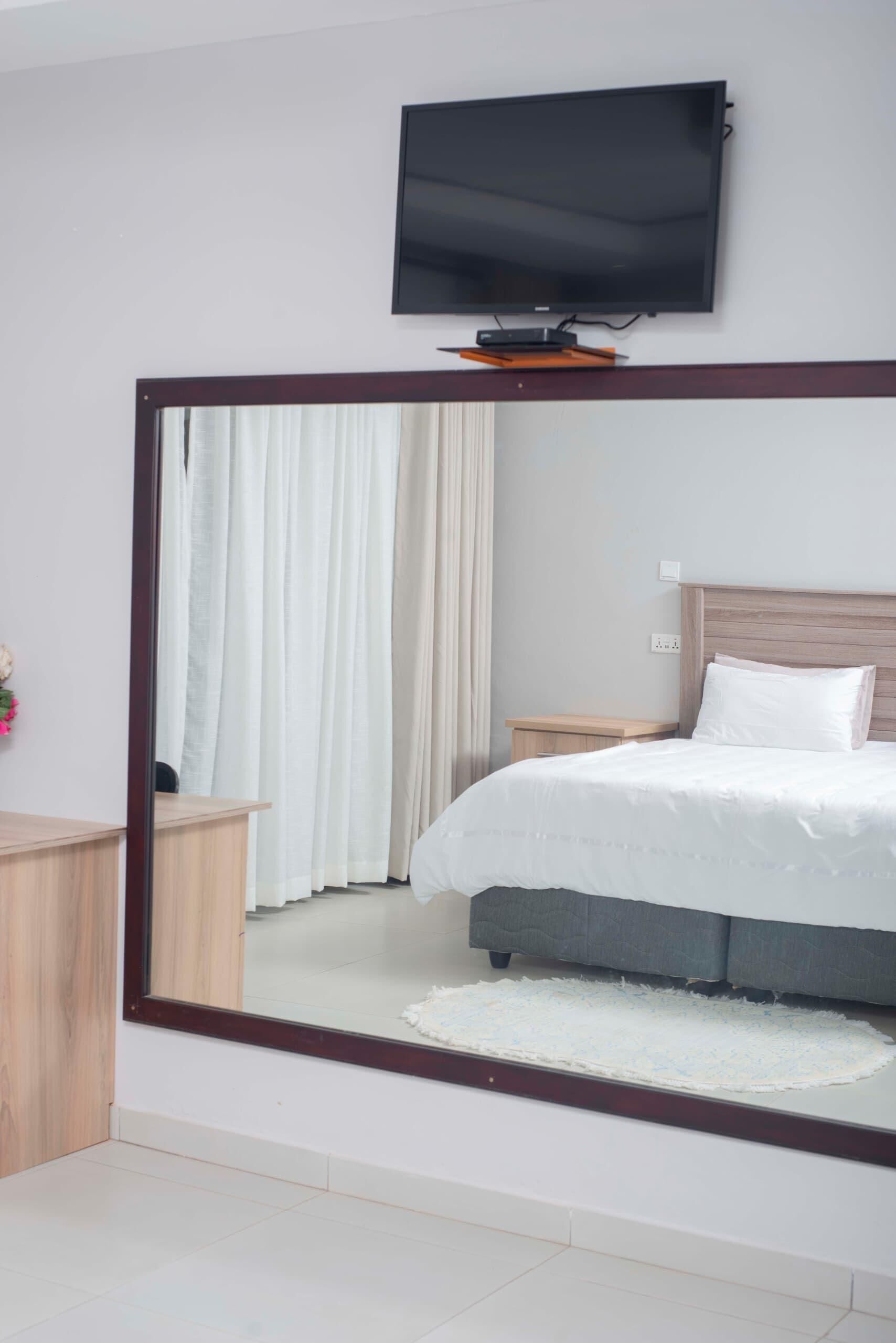 A bedroom with a large mirror, a bed with white bedding, a wooden nightstand, a TV mounted on the wall, and a window with curtains.