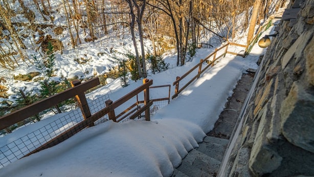 A snow-covered staircase with wooden railings and stone walls, surrounded by trees and a fence.