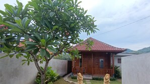 A wooden house with a tiled roof, a small garden with a tree and two statues, and a stone pathway leading to the entrance. - Saghara Villa Amed (Karangasem)