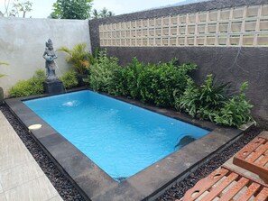 A rectangular swimming pool with a statue, surrounded by lush greenery and a textured wall. - Saghara Villa Amed (Karangasem)
