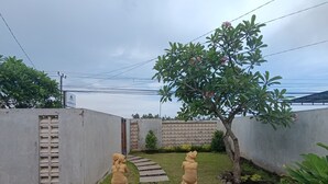 A garden with two stone statues, a concrete wall, and a tree with pink flowers. - Saghara Villa Amed (Karangasem)