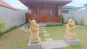 A wooden house with a tiled roof, a porch, and two statues in the garden. - Saghara Villa Amed (Karangasem)