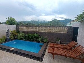 A swimming pool with a statue, lounge chairs, and a view of green hills. - Saghara Villa Amed (Karangasem)