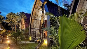 A well-lit garden area with a pathway, palm trees, and a building with a unique architectural design. - The Suwi Huts (Penida Island)
