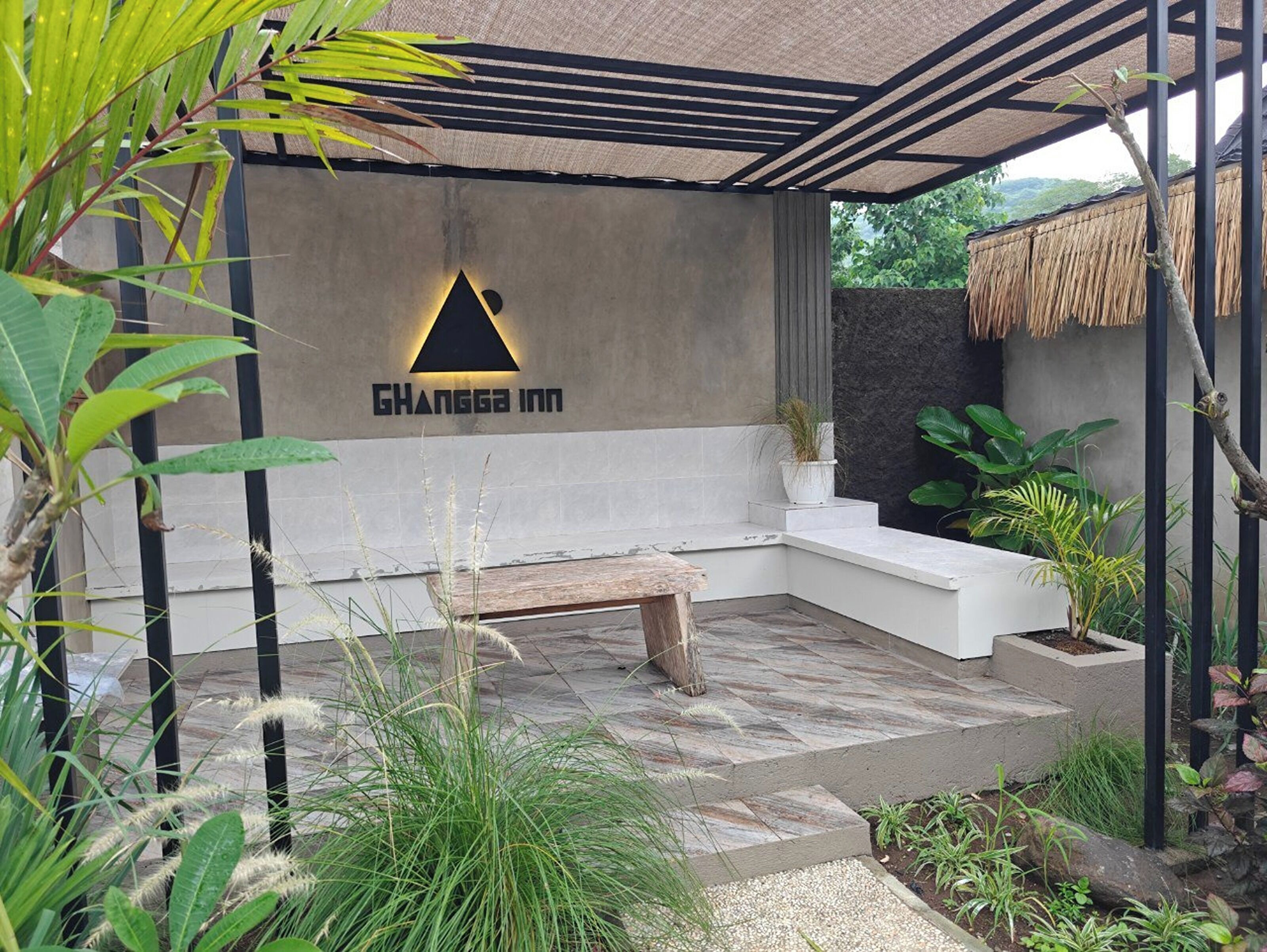 Un área de estar moderna al aire libre, con un banco, el logotipo sujeto a la pared y una estructura de techo de paja.