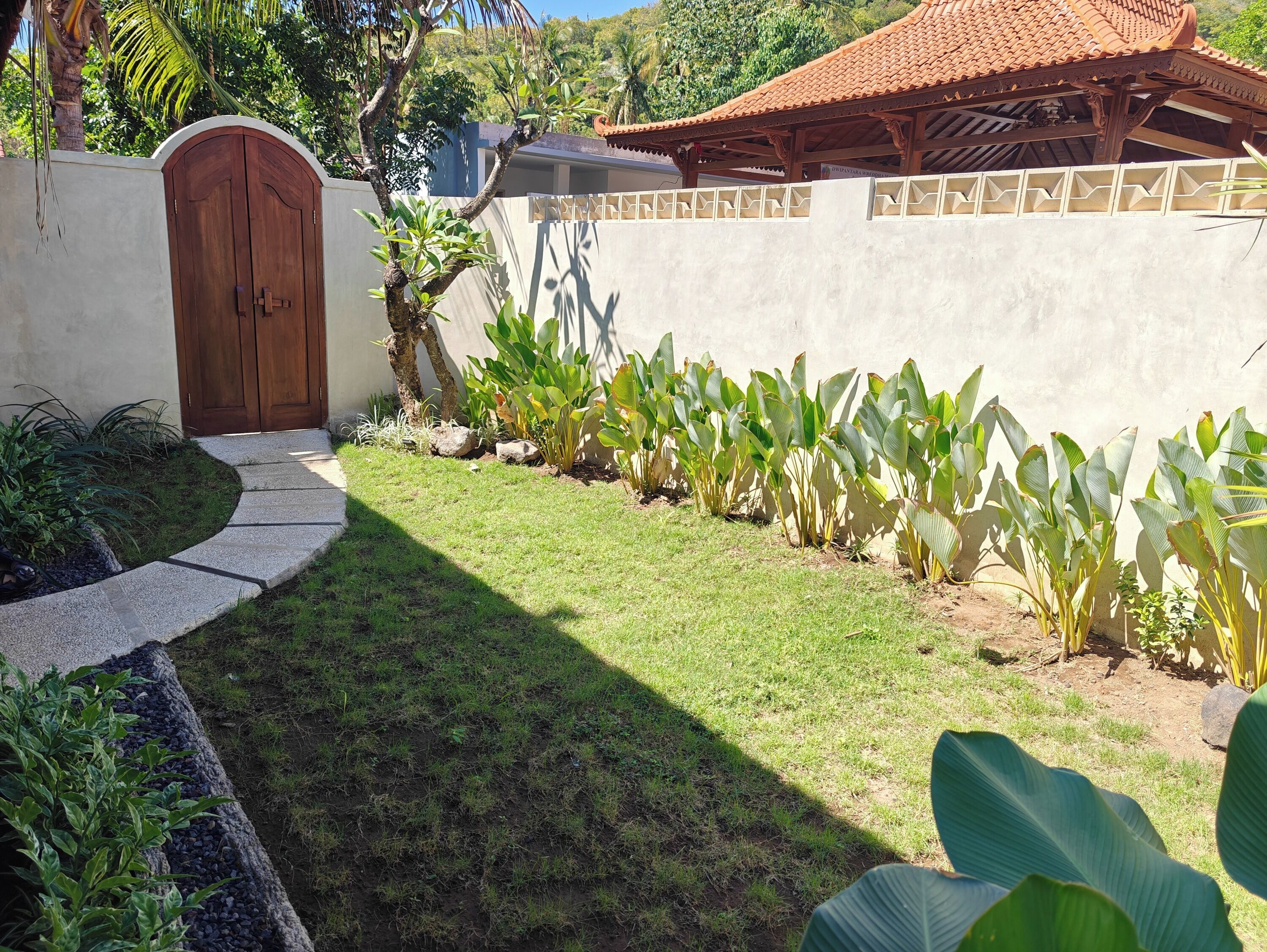 A well-maintained garden with a paved pathway leading to a wooden door, surrounded by lush greenery and a tiled roof structure.