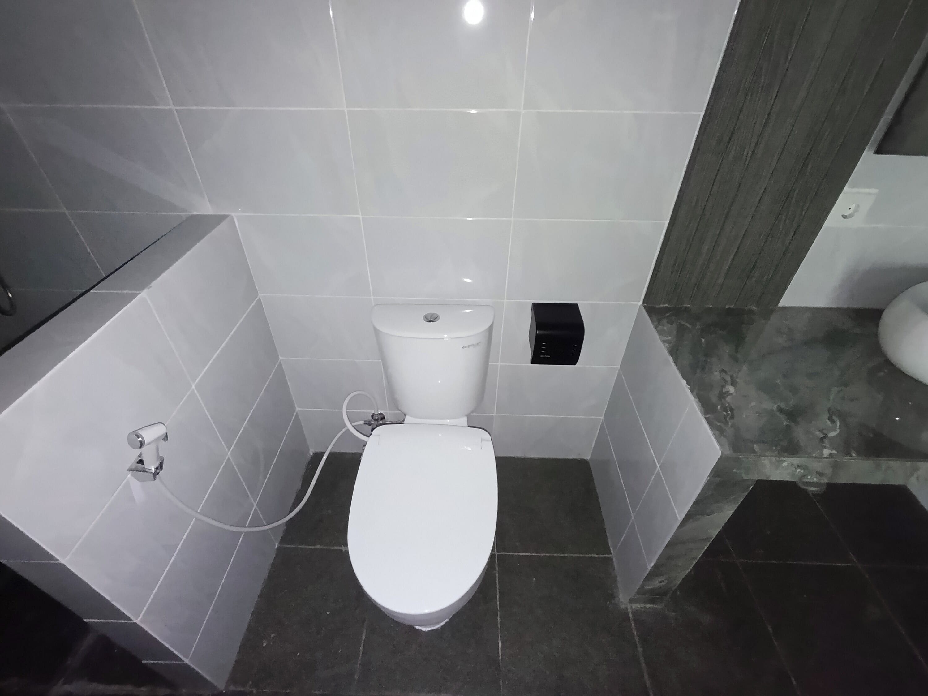 A small, modern bathroom with a white toilet, a hand dryer, and a soap dispenser. The floor is tiled with a mix of light and dark tiles.