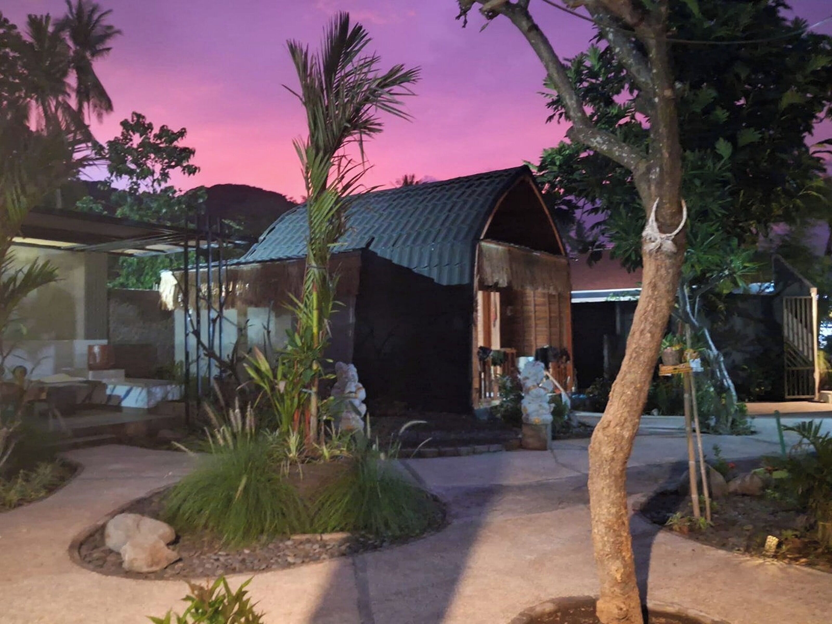 A rustic building with a thatched roof, surrounded by tropical vegetation and a paved pathway.
