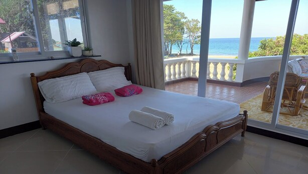 A neatly made bed with white linens and pink pillows, a wooden headboard, and a view of the sea through large windows. - Seven Waves Beachfront (Bauang)