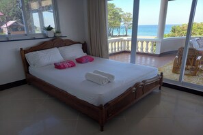 Una cama bien hecha con sábanas blancas y almohadas rosas, un cabecero de madera y vistas al mar a través de amplios ventanales.