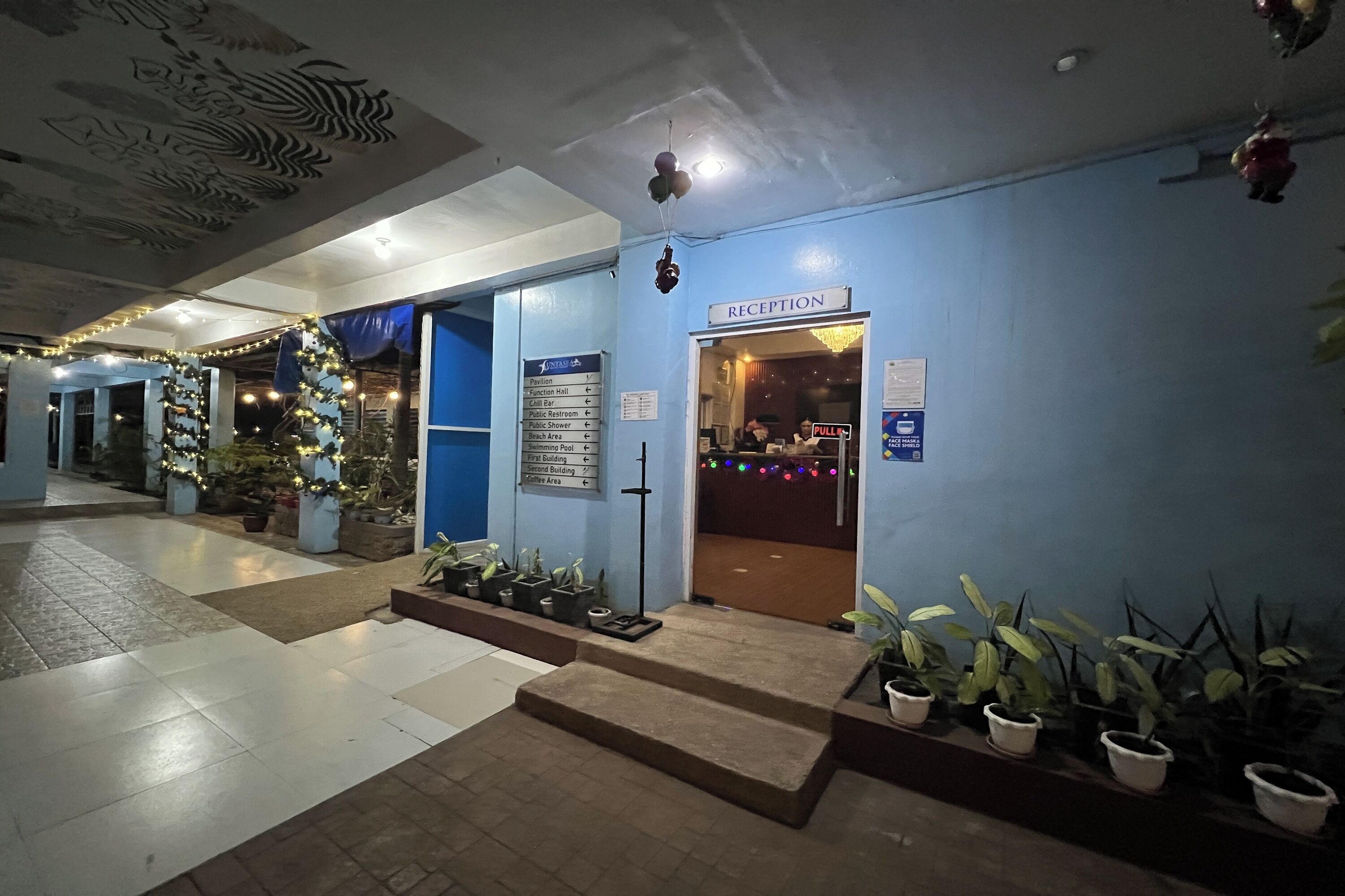 A hotel reception area with a blue wall, a reception desk, and a sign indicating the reception. There are potted plants and a staircase leading to the entrance.